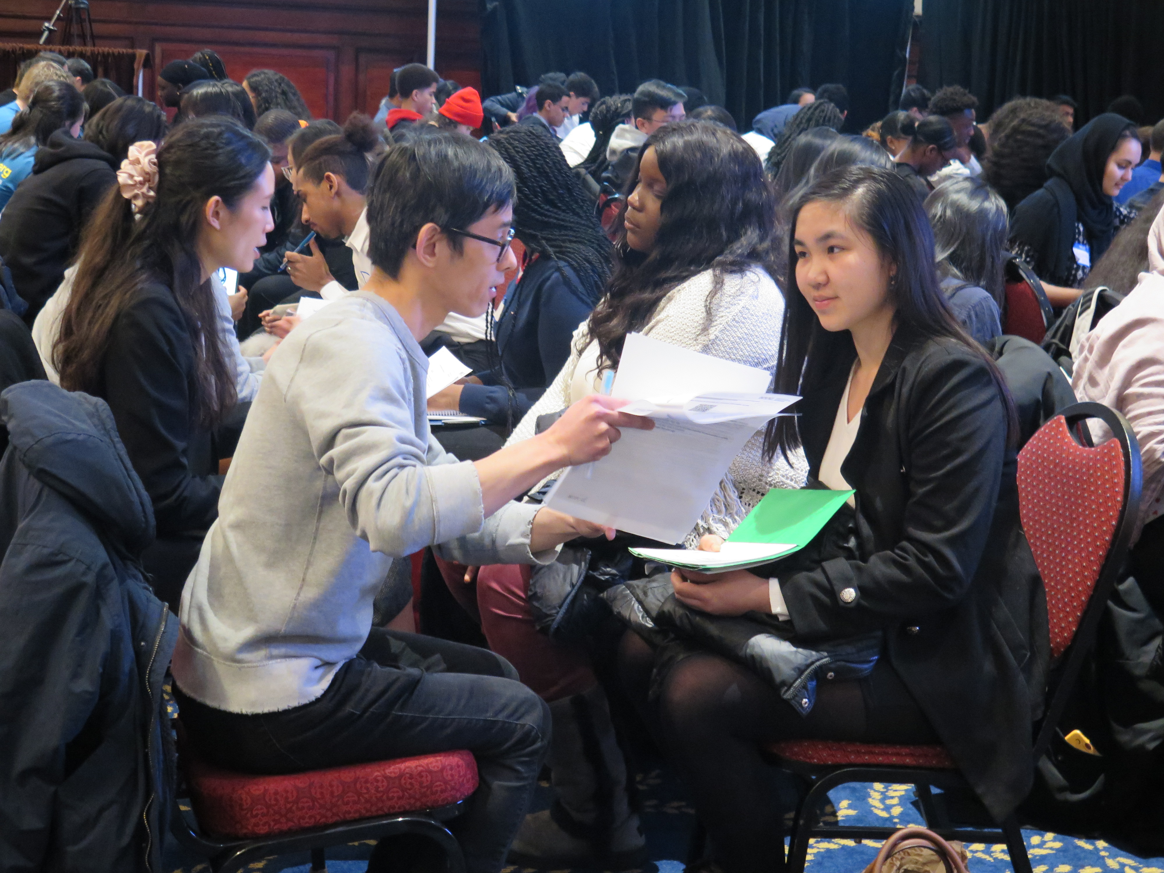 A Ladders for Leaders internship training workshop. A large room full of chairs with groups of two sitting across from each other. In the foreground, a man is speaking to a young woman, and holding up a few pieces of paper.